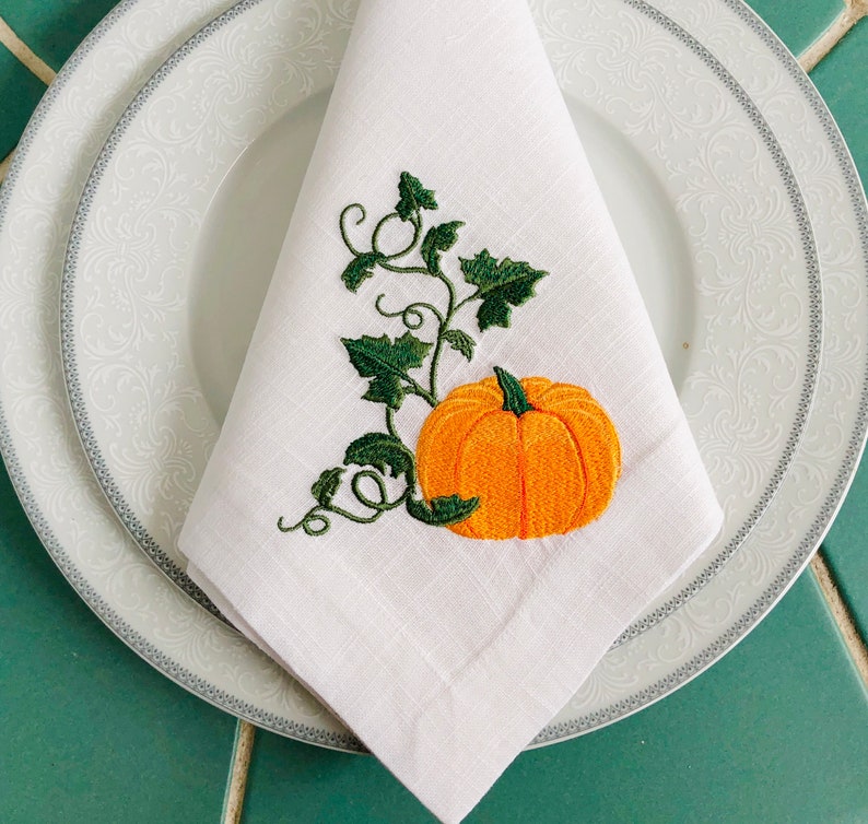 May include: White linen napkin featuring an embroidered pumpkin and vine design. The pumpkin is orange with a green stem and leaves. The vine is green with leaves.