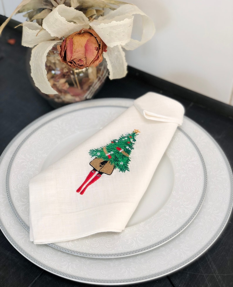 May include: White linen napkin with a red and green embroidered design of a person carrying a Christmas tree.