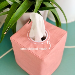Puede incluir: Una funda para caja de pañuelos de lino rosa con un círculo bordado blanco alrededor de la abertura. El texto "embroidered mouth" está escrito debajo del círculo.