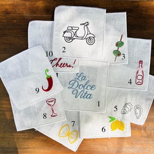 May include: A collection of white linen handkerchiefs, each embroidered with a different design. Designs include a scooter, olives, a bottle of wine, oysters, a lemon, pasta, a cocktail glass, a chili pepper, and the phrase "Cheers!"