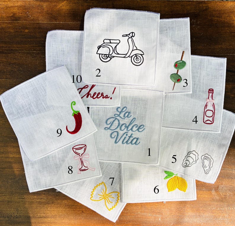 May include: A collection of white linen handkerchiefs, each embroidered with a different design. Designs include a scooter, olives, a bottle of wine, oysters, a lemon, pasta, a cocktail glass, a chili pepper, and the phrase "Cheers!"