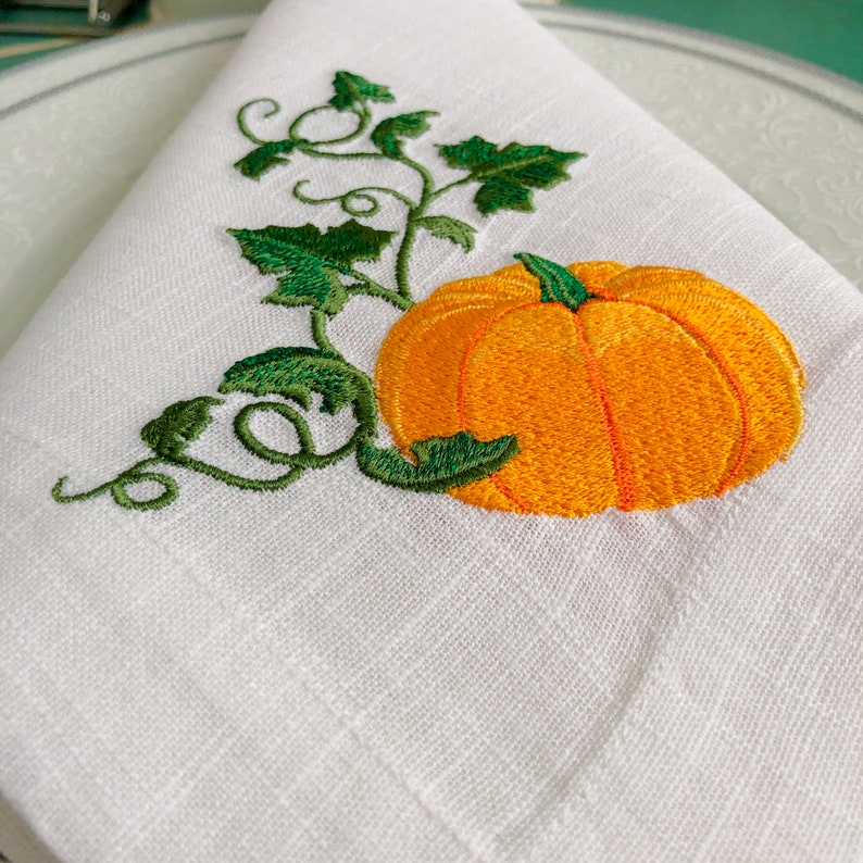 May include: White linen napkin with an embroidered orange pumpkin and green vine. The pumpkin has a stem and is surrounded by leaves.