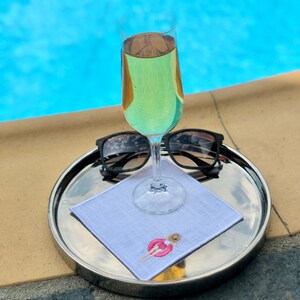 May include: A silver tray with a white linen napkin, a glass of white wine, and a pair of sunglasses. The napkin has an embroidered design of a person in a life preserver.