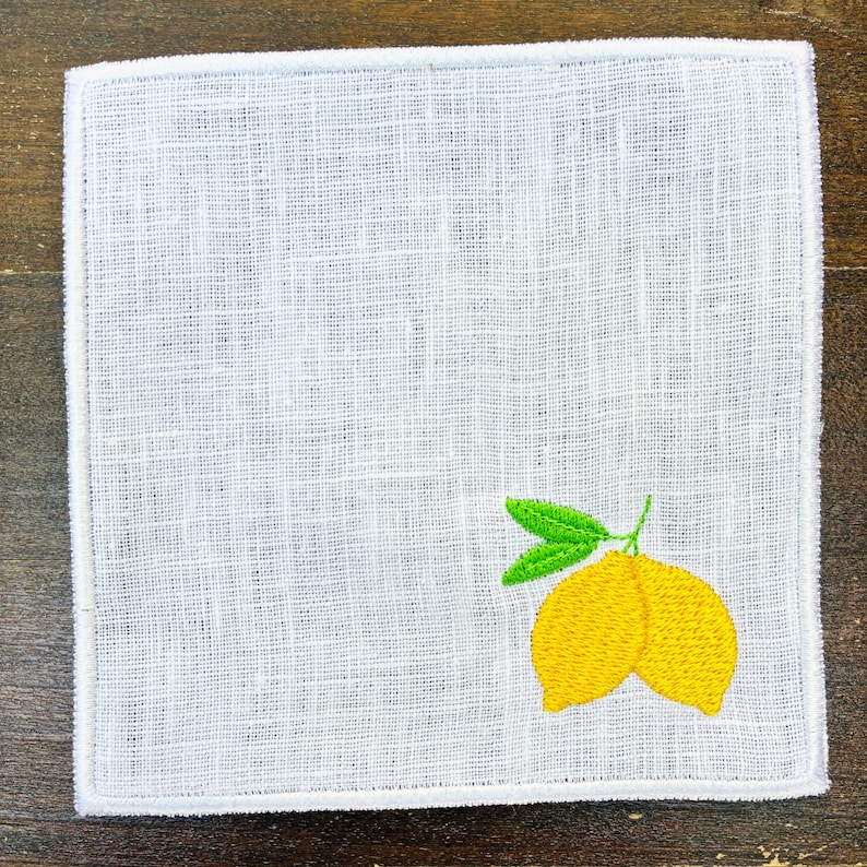 May include: White square linen napkin with a stitched lemon design in yellow and green. The napkin has a white border and a textured weave.