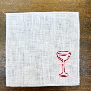 May include: Square white linen handkerchief with a red wine glass embroidered on the corner. The wine glass has a pink bow and ribbon. The handkerchief is on a wooden surface.