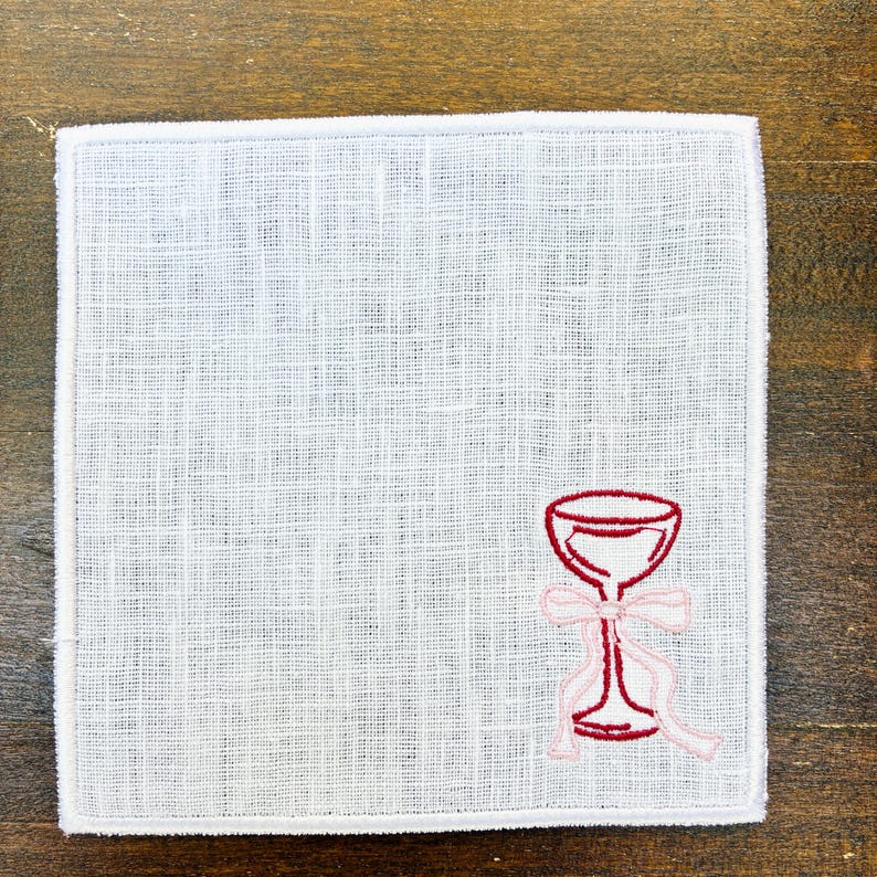 May include: Square white linen handkerchief with a red wine glass embroidered on the corner. The wine glass has a pink bow and ribbon. The handkerchief is on a wooden surface.
