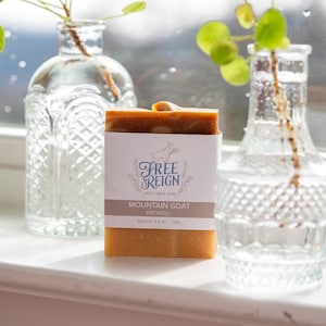 May include: A bar of Mountain Goat Patchouli soap with a white label featuring a logo and text. The soap is approximately 4.5 oz (128g). Two clear glass vases with green plants are in the background.