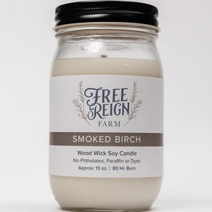 May include: A glass jar candle with a black lid and a white label. The label says "Free Reign Farm" and "Smoked Birch". The candle is a wood wick soy candle with no phthalates, paraffin, or dyes. It is approximately 13 ounces and has an 80-hour burn time.