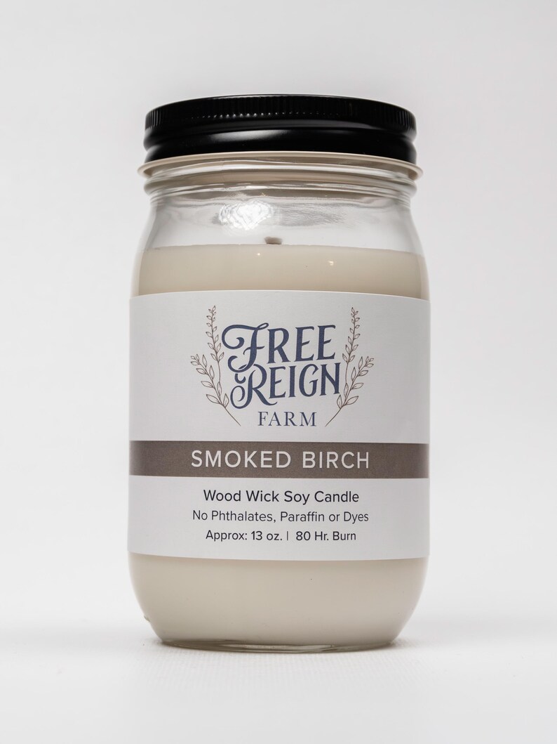 May include: A glass jar candle with a black lid and a white label. The label says "Free Reign Farm" and "Smoked Birch". The candle is a wood wick soy candle with no phthalates, paraffin, or dyes. It is approximately 13 ounces and has an 80-hour burn time.