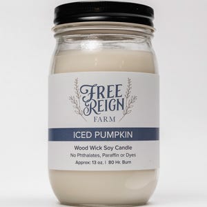 May include: A glass jar candle with a black lid and a white label. The label says "FREE REIGN FARM" and "ICED PUMPKIN". The candle is a wood wick soy candle with no phthalates, paraffin or dyes. It is approximately 13 ounces and has an 80 hour burn time.