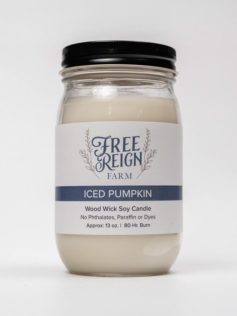 May include: A glass jar candle with a black lid and a white label. The label says "FREE REIGN FARM" and "ICED PUMPKIN". The candle is a wood wick soy candle with no phthalates, paraffin or dyes. It is approximately 13 ounces and has an 80 hour burn time.