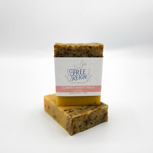 May include: Two bars of yellow goat milk soap with brown flecks. The top bar has a white label with the text "Free Reign Goat Milk Soap" and "Summer Night Walk Lavender Approx. 4.5 oz. (128g)".