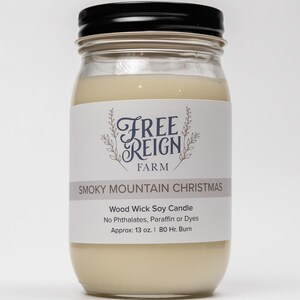 May include: A glass jar candle with a black lid. The label reads "FREE REIGN FARM" and "SMOKY MOUNTAIN CHRISTMAS". The candle is a wood wick soy candle with no phthalates, paraffin, or dyes. It is approximately 13 ounces and has an 80-hour burn time.