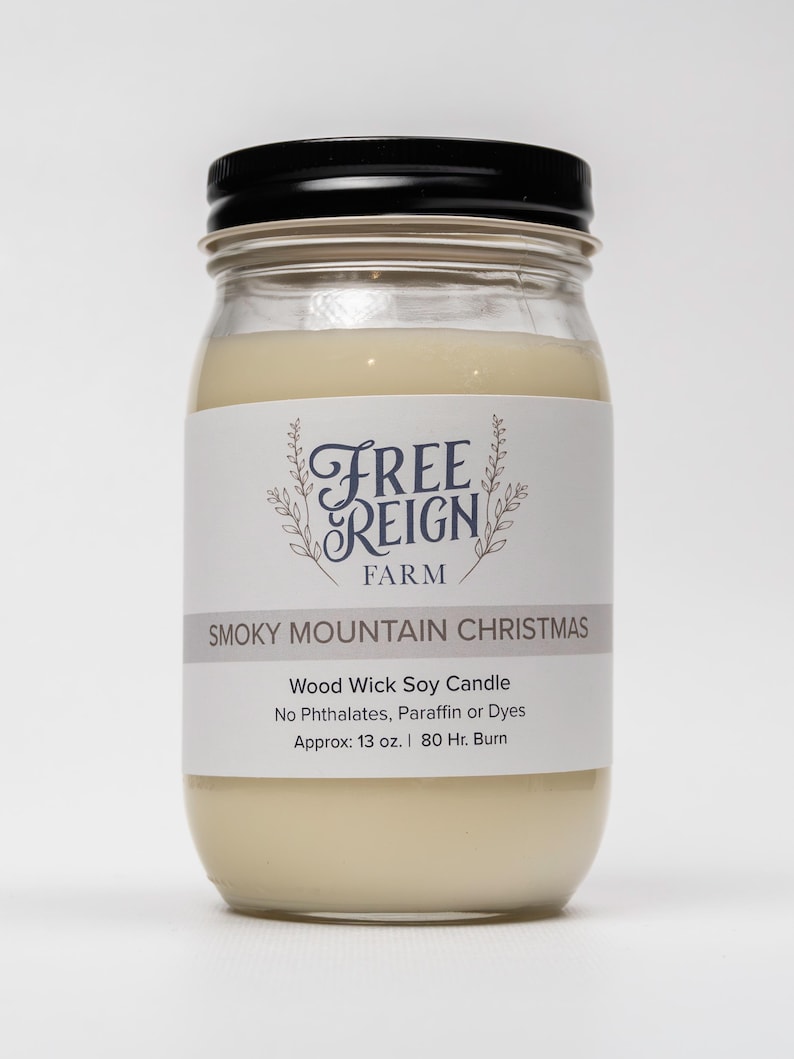May include: A glass jar candle with a black lid. The label reads "FREE REIGN FARM" and "SMOKY MOUNTAIN CHRISTMAS". The candle is a wood wick soy candle with no phthalates, paraffin, or dyes. It is approximately 13 ounces and has an 80-hour burn time.