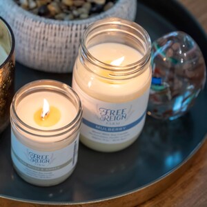 May include: Two glass jars with lit candles inside. The jars are labeled "Free Reign Farm" and "Mulberry".