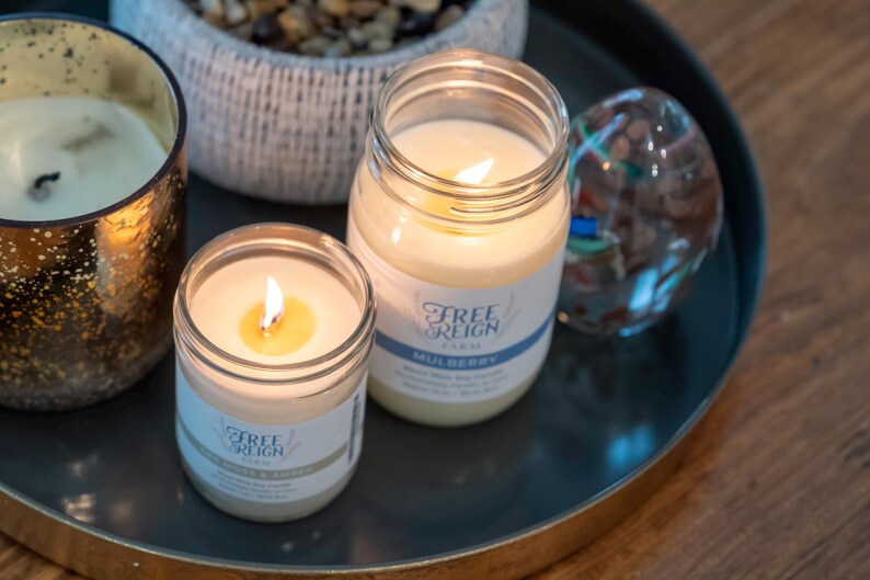 May include: Two glass jars with lit candles inside. The jars are labeled "Free Reign Farm" and "Mulberry".