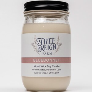 May include: A glass jar candle with a black lid. The label reads "FREE REIGN FARM" in a decorative font and "BLUEBONNET" in a pink rectangle. The candle is a wood wick soy candle with no phthalates, paraffin or dyes. It is approximately 13 ounces and has an 80 hour burn time.