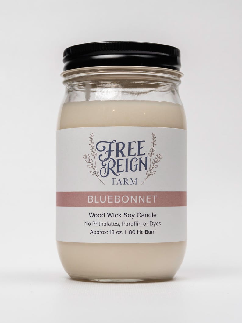 May include: A glass jar candle with a black lid. The label reads "FREE REIGN FARM" in a decorative font and "BLUEBONNET" in a pink rectangle. The candle is a wood wick soy candle with no phthalates, paraffin or dyes. It is approximately 13 ounces and has an 80 hour burn time.