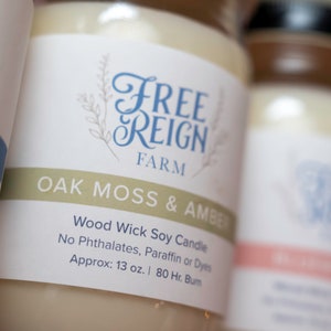 May include: A white jar candle with a label that reads "FREE REIGN FARM OAK MOSS & AMBER Wood Wick Soy Candle No Phthalates, Paraffin or Dyes Approx: 13 oz. | 80 Hr. Burn"