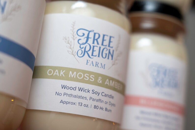 May include: A white jar candle with a label that reads "FREE REIGN FARM OAK MOSS & AMBER Wood Wick Soy Candle No Phthalates, Paraffin or Dyes Approx: 13 oz. | 80 Hr. Burn"