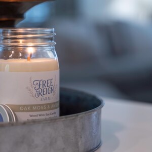 May include: A glass jar candle with a wood wick, labeled "Free Reign Farm Oak Moss & Amber" Wood Wick Soy Candle. The candle is lit and sitting on a metal tray.