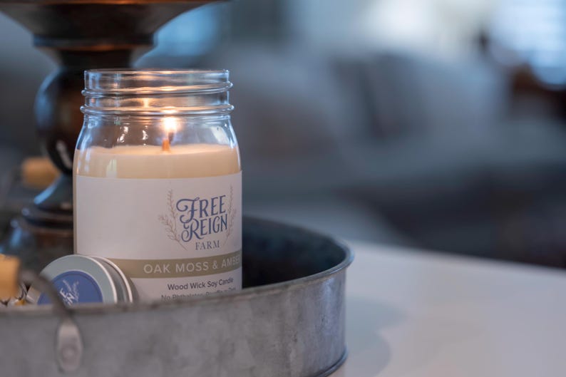 May include: A glass jar candle with a wood wick, labeled "Free Reign Farm Oak Moss & Amber" Wood Wick Soy Candle. The candle is lit and sitting on a metal tray.