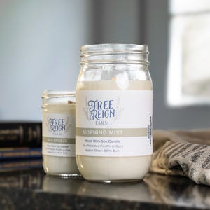 May include: Two glass jars with white labels and black text. The labels read "Free Reign Farm" and "Morning Mist". The jars contain soy candles with wooden wicks. The candles are labeled "Wood Wick Soy Candle" and "Free Reign Farm".