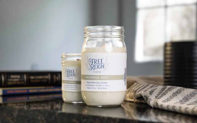 May include: Two glass jars with white labels and black text. The labels read "Free Reign Farm" and "Morning Mist". The jars contain soy candles with wooden wicks. The candles are labeled "Wood Wick Soy Candle" and "Free Reign Farm".