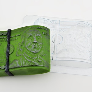 May include: A green plastic mold of a stack of $100 bills with a black rubber band. The mold is designed to create a realistic-looking stack of money for crafts or decorations.