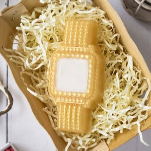 May include: A gold-colored soap bar shaped like a wristwatch. The soap has a white square in the center, resembling a watch face.