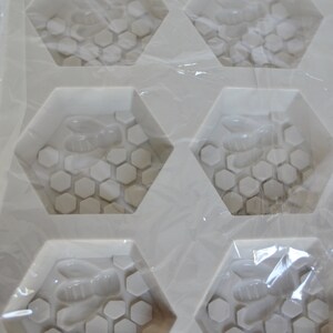 May include: A white silicone mould with six hexagonal cavities. Each cavity features a bee and honeycomb design. The mould is wrapped in clear plastic, suggesting it's for crafting soaps or similar items.