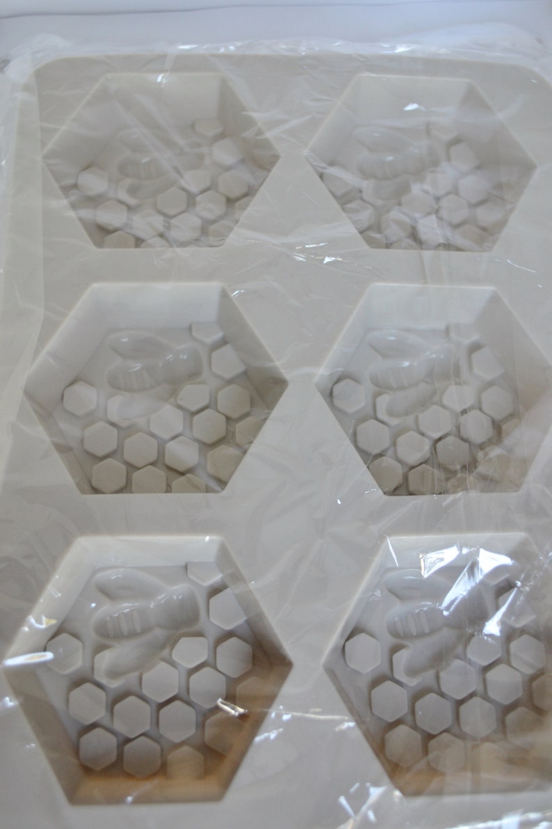 May include: A white silicone mould with six hexagonal cavities. Each cavity features a bee and honeycomb design. The mould is wrapped in clear plastic, suggesting it's for crafting soaps or similar items.