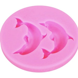 May include: Pink silicone mold with two dolphin shapes. The mold is designed for making cake decorations or other edible treats.