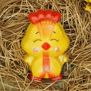 Può includere: Una bomba da bagno a forma di pulcino giallo con accenti rossi e arancioni. Il pulcino ha un viso sorridente con gli occhi chiusi e un becco rosso. La bomba da bagno è seduta in un nido di paglia.