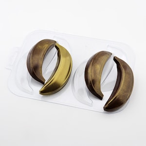 May include: A clear plastic mold containing two pairs of curved, banana-shaped cavities. Each cavity features a metallic bronze and dark brown finish, creating a realistic, shaded appearance for the molded shapes within the transparent tray.