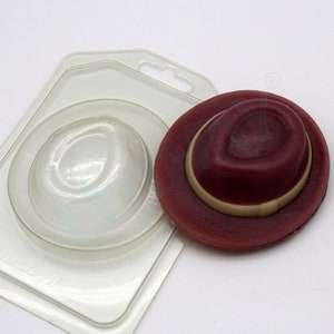 Könnte beinhalten: Eine transparente Plastikform zum Herstellen eines roten Fedora-Hutes. Die Form wird neben einem fertigen roten Fedora-Hut mit einem cremefarbenen Band gezeigt.