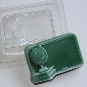 May include: A green soap in the shape of a chalkboard, featuring a globe and books, alongside a clear plastic mould of the same design. The soap has a white outline and is on a white surface.