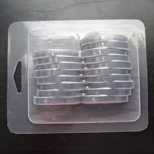 May include: A clear plastic packaging tray containing two stacks of gray, round, silicone molds. The molds are stacked on top of each other and are designed for making candles or soap.