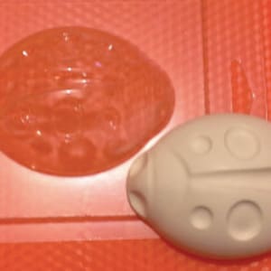 Könnte beinhalten: Eine weiße Marienkäfer-Form mit einer strukturierten Oberfläche und fünf erhabenen Kreisen. Die Form liegt neben einer durchsichtigen Plastikform mit dem gleichen Design.