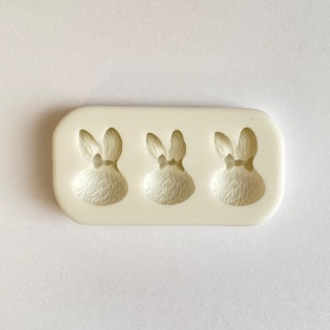 May include: White silicone mold with three bunny head cavities. Each bunny head has a bow tie and fluffy fur texture.