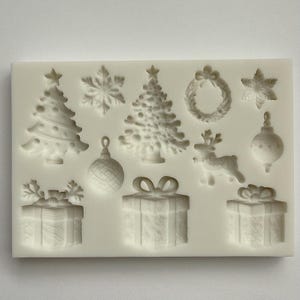May include: A white silicone mold featuring Christmas-themed shapes. The mold includes a Christmas tree, snowflake, wreath, star, ornament, reindeer, and gift box designs. Perfect for crafting festive decorations or treats.