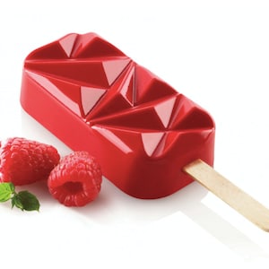 Może przedstawiać: Czerwona forma do lodów na patyku z geometrycznym wzorem i drewnianym patyczkiem. Forma leży na białym tle z dwoma czerwonymi malinami i zielonym listkiem.
