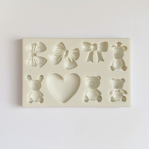 Może przedstawiać: Forma silikonowa z siedmioma wnękami do tworzenia dekoracji z lukru lub gum paste. Forma zawiera serce, trzy kokardki i trzy pluszowe misie.