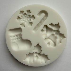 Puede incluir: Un molde de silicona blanco con diseños navideños. El molde presenta un copo de nieve, un bastón de caramelo, un calcetín, acebo y campanas. El molde circular está diseñado para crear decoraciones o golosinas festivas.