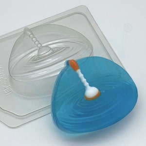 May include: A clear plastic mold with a rippled surface, containing a blue, teardrop-shaped object with a white and orange spoon-like handle. Another clear mold is visible in the background.