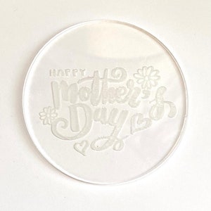 Può includere: Timbro in acrilico circolare bianco con il testo "Happy Mother's Day" in un carattere corsivo con un design a forma di cuore e fiore.