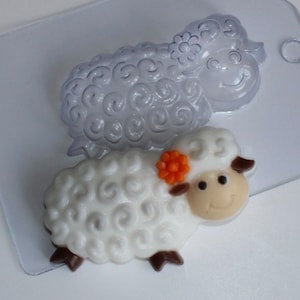 May include: A white chocolate sheep with a brown nose and hooves, a small orange flower, and a curly white coat. The sheep is standing on a white surface. There is a clear plastic mold of a sheep in the background.