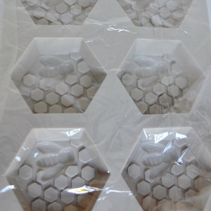 May include: A white silicone mould with six hexagonal cavities. Each cavity features a bee and honeycomb design. The mould is designed for soap making or crafting.
