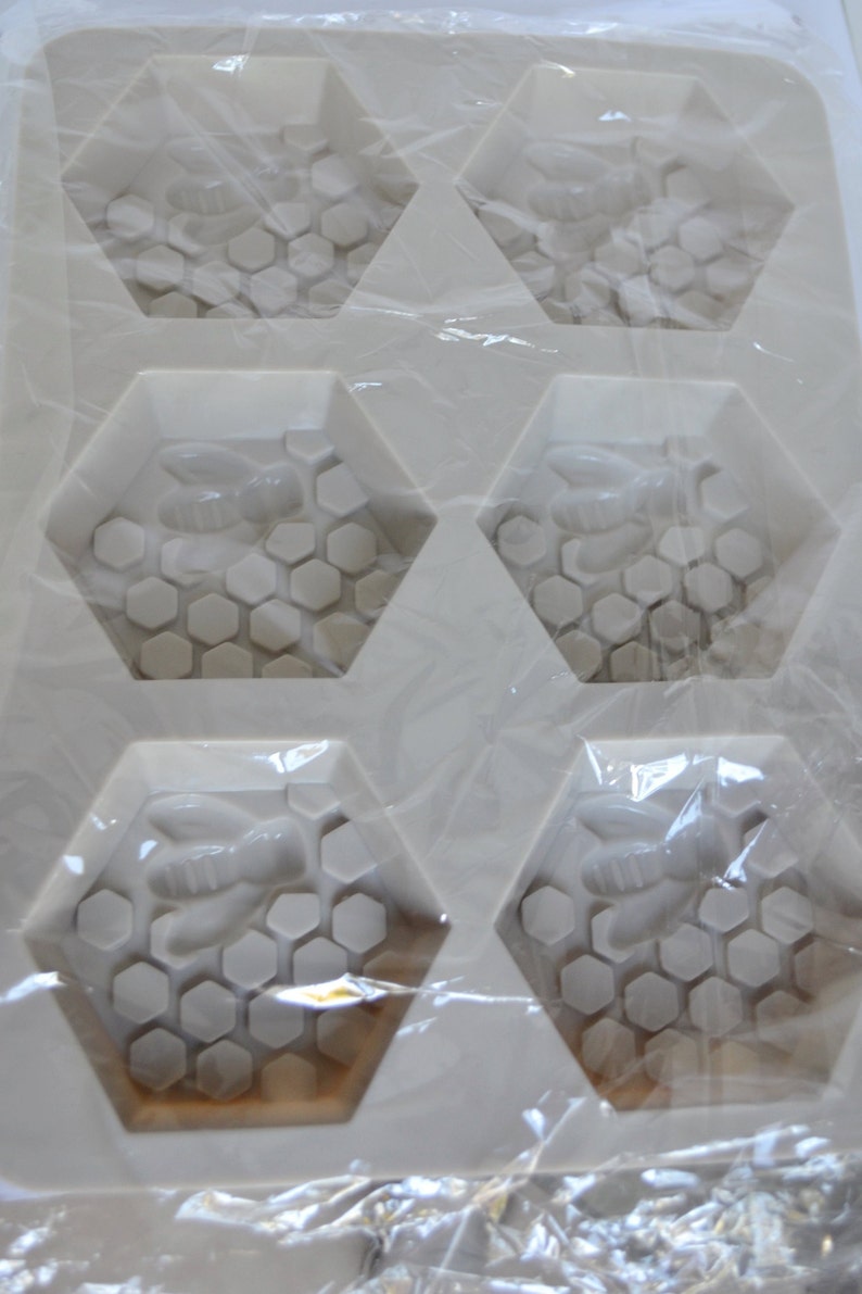 May include: A white silicone mould with six hexagonal cavities. Each cavity features a bee and honeycomb design. The mould is designed for soap making or crafting.