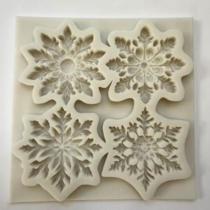 May include: A square, off-white silicone mold featuring four snowflake designs. Each snowflake has a detailed, symmetrical pattern with intricate leaf-like details. The mold is designed for creating decorative elements.